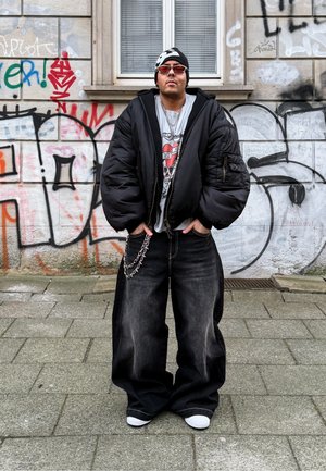 Una persona con una chaqueta acolchada negra sobre una camiseta gráfica, vaqueros negros holgados con una cadena y zapatillas blancas, de pie frente a un graffiti.