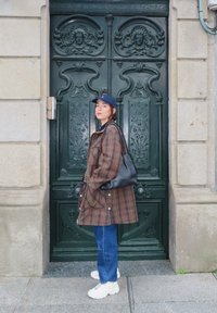 Casaco oversized em padrão xadrez castanho, jeans azuis, sapatilhas brancas, bolsa de ombro preta, boné azul-marinho; em pé ao lado de uma porta verde detalhada com design ornamental.