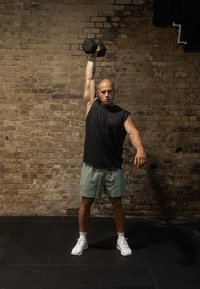 Μαύρο αμάνικο μπλουζάκι, ανοιχτό πράσινο σορτς και άσπρα παπούτσια; άνδρας σηκώνει ένα μαύρο αλτήρα πάνω από το κεφάλι του με φόντο έναν τοίχο από τούβλα.