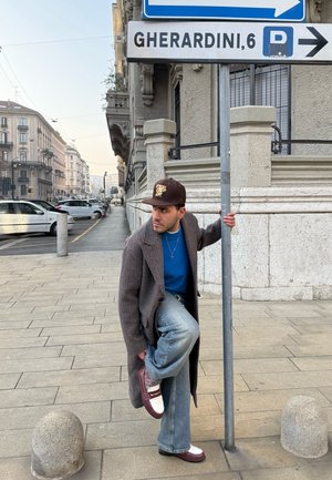 Hombre con abrigo largo y gorra atándose los cordones mientras sostiene un poste con señales junto a una calle con coches estacionados y edificios.