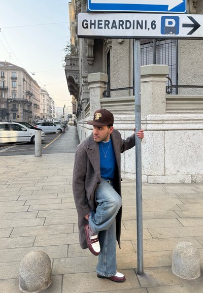 Hombre con abrigo largo y gorra atándose los cordones mientras sostiene un poste con señales junto a una calle con coches estacionados y edificios.