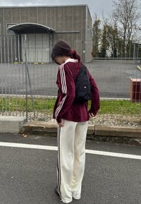 Maronjakk med hvite striper, hvite bukser med striper på siden og en svart ryggsekk. Antrekket har en avslappet passform og et teksturert stoff.