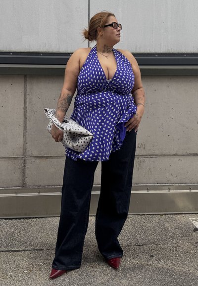 Mujer con tatuajes que lleva un top halter azul con lunares, pantalones anchos de pierna oscuros, zapatos puntiagudos rojos, gafas de sol negras y sostiene un clutch blanco con estampado.