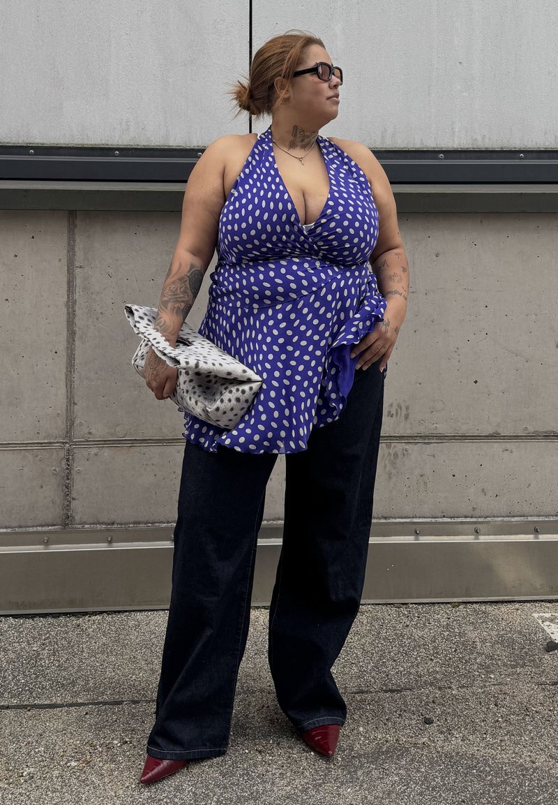Woman with tattoos wears a blue polka-dot halter top, dark wide-leg pants, red pointed shoes, black sunglasses, and holds a white patterned clutch.