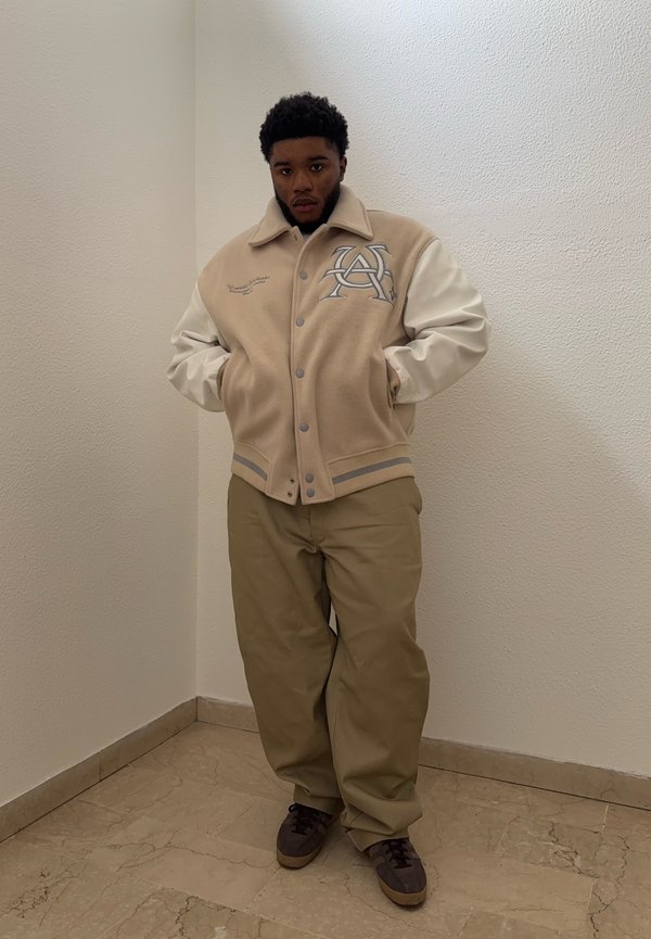 Veste de baseball beige avec des manches en cuir blanc et un logo, associée à un pantalon kaki et des baskets brunes sur un sol carrelé.