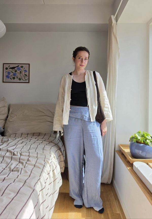Femme debout dans une chambre portant une veste légère, un haut noir, un pantalon bleu et tenant un sac marron près d'une fenêtre avec une plante en pot.