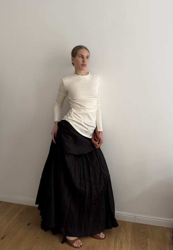 Cremefarvet langærmet top med rynkede detaljer, parret med en flydende sort maxinederdel i en tekstureret stof og plisseret design.