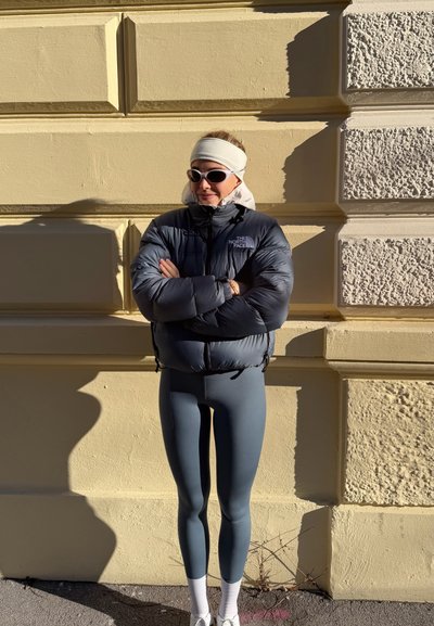 Mujer vestida con chaqueta acolchada gris, leggings azul claro, zapatillas blancas, gafas de sol y cinta blanca en la cabeza, de pie contra una pared texturizada amarilla bajo la luz del sol.