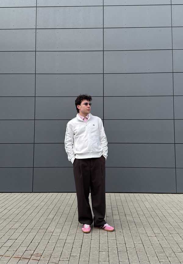 Moški z belim jopičem, roza srajco, temnimi širokimi hlačami in roza čevlji stoji z rokami v žepih ob sivi obloženi steni.
