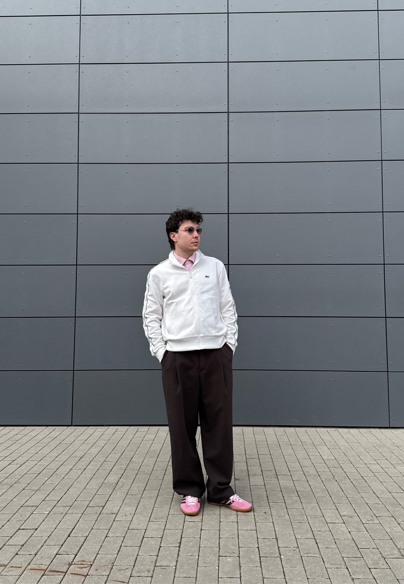 Homme portant une veste blanche, une chemise rose, un pantalon large foncé et des chaussures roses, debout les mains dans les poches devant un mur carrelé gris.