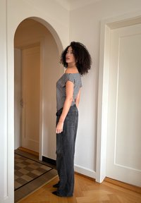 Haut rayé à épaules dénudées en noir et blanc, associé à un jean noir taille haute à jambes larges. Modèle debout dans un espace intérieur neutre.