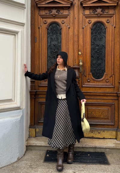 Mujer con abrigo negro, falda a cuadros, gorro negro y botas marrones, apoyada con una mano en una pared blanca y sosteniendo un bolso amarillo claro junto a una gran puerta de madera.