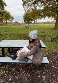 Pilka megzta kepurė, ruda dirbtinio kailio striukė, šviesiai pilkos kelnės, juodi vandeniui atsparūs batai. Sėdi prie pikniko stalo su natūraliu fonu.