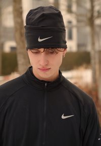 Jeune homme portant un bonnet noir Nike et une veste, regardant vers le bas, en plein air avec des arbres et des bâtiments flous en arrière-plan.