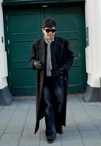 Manteau noir, écharpe à rayures horizontales, gants noirs, jeans foncés et chaussures noires brillantes devant une porte verte. Cadre urbain.