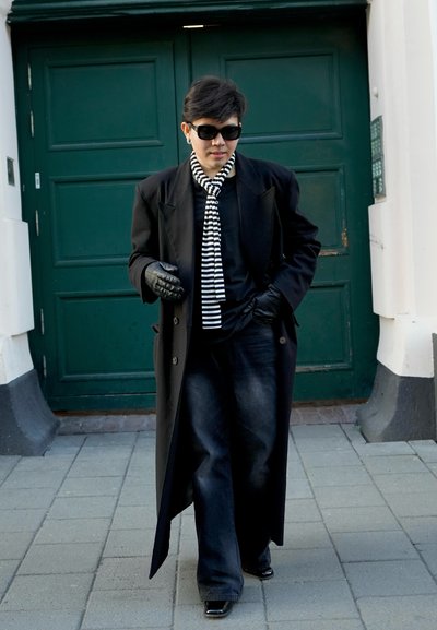 Abrigo negro, bufanda de rayas horizontales, guantes negros, vaqueros oscuros y zapatos negros brillantes frente a una puerta verde. Escenario urbano en la calle.