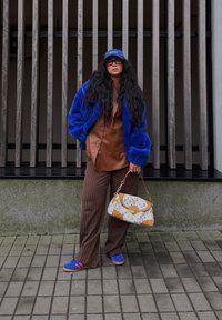 Giacca blu soffice, camicia in pelle marrone, pantaloni marrone a righe, sneakers blu e una borsa beige con motivo. In piedi contro una recinzione.