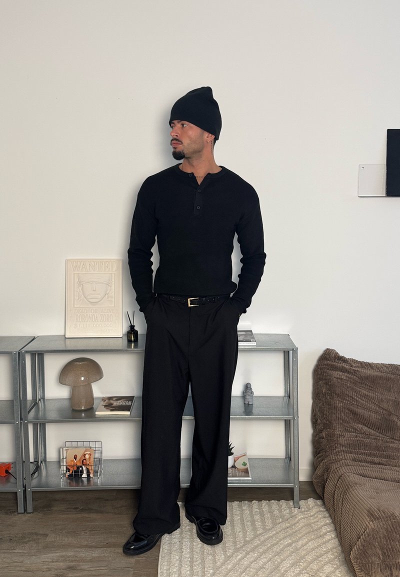 Chemise henley à manches longues noire, pantalon noir et bonnet noir. La tenue se caractérise par une coupe décontractée et un design minimaliste.