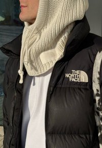 Personne portant une capuche en tricot crème avec cordons de serrage et une veste matelassée noire The North Face par-dessus une chemise blanche.