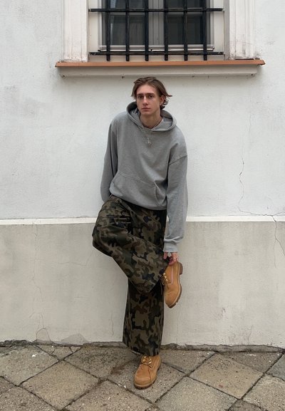 Sudadera gris con capucha, pantalones de camuflaje holgados y botas beige. El sujeto se apoya contra una pared con una ventana con barrotes al fondo.