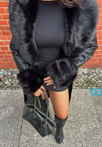 Vrouw in een zwart kunstbont gevoerd leren jas, zwarte jurk, laarzen tot de knie, met een zwarte leren handtas, staande op een stenen stoep.