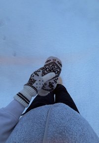 Personne portant des moufles à motifs et des vêtements chauds tient une main gantée contre le sol enneigé, vue de dessus.