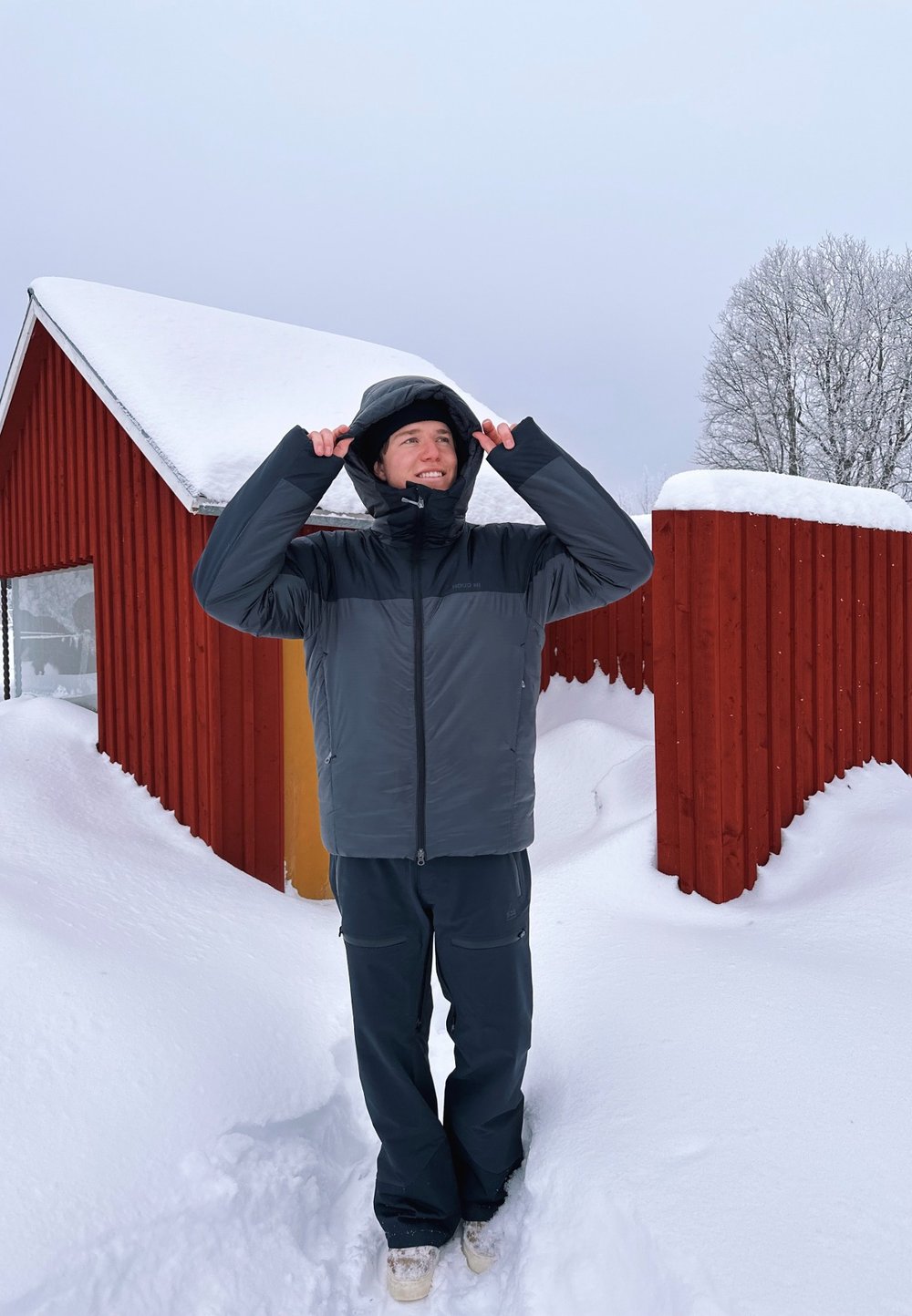 Grijze pufferjack met een capuchon, met een gladde textuur en een ritssluiting. Gepaard met donkere broeken, tegen een besneeuwd landschap.