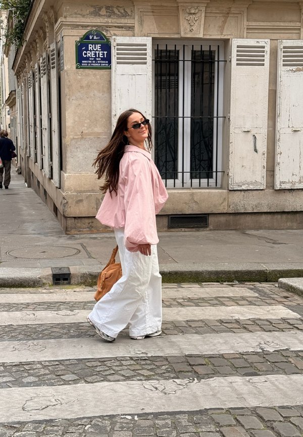 Femme portant une veste rose et un pantalon blanc marchant sur un passage piéton pavé devant un vieux bâtiment avec des volets fermés.