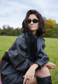 Jachetă oversized din piele neagră cu guler înalt, asortată cu ochelari de soare negri. O geantă cu accente aurii este sprijinită pe umăr.