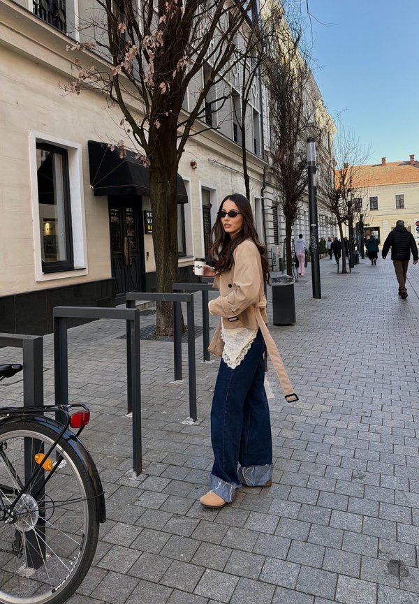 Žena v slnečných okuliaroch a béžovom kabáte stojí na mestskom chodníku, drží šálku kávy pri stojanoch na bicykle a holých stromoch.