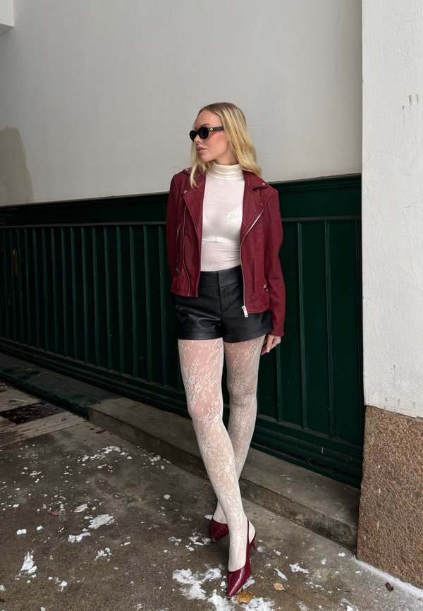 Szőke nő napszemüvegben, piros dzsekiben, fehér garbóban, fekete rövidnadrágban, mintás fehér harisnyában és piros magassarkú cipőben, zöld-fehér fal előtt állva.