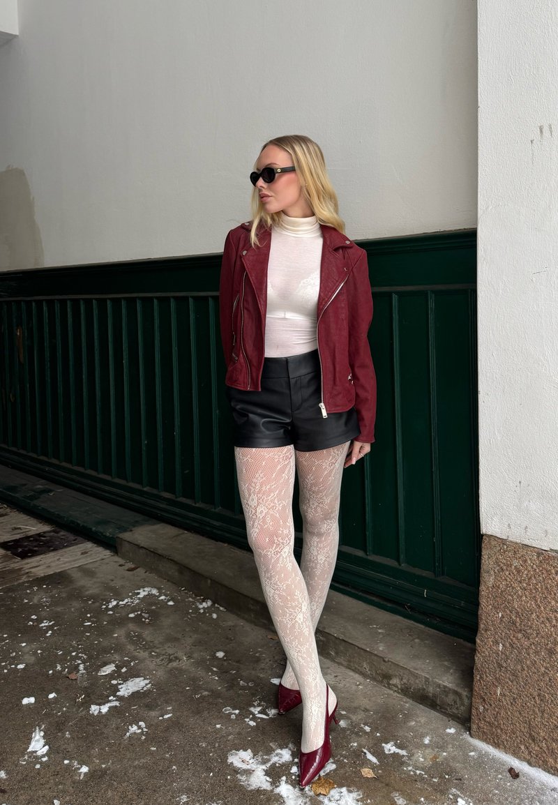 Blond kvinne med solbriller, rød jakke, hvit høyhalset genser, svarte shorts, mønstrede hvite strømpebukser og røde hæler som står foran en grønn og hvit vegg.