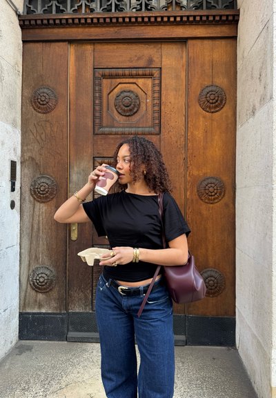 Mujer con cabello rizado bebiendo café, sosteniendo una bandeja con taza, de pie frente a una gran puerta de madera ornamentada.