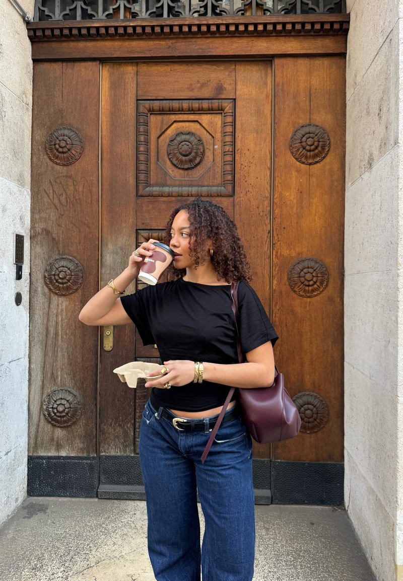 Femme aux cheveux bouclés buvant du café, tenant un plateau avec une tasse, debout devant une grande porte en bois ornée.
