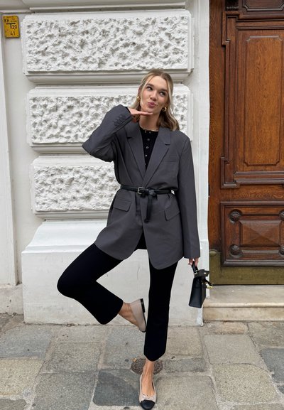 Blazer gris de gran tamaño con cintura ceñida por cinturón, combinado con pantalones negros de campana y bailarinas beige con puntera negra. Sosteniendo un bolso negro.
