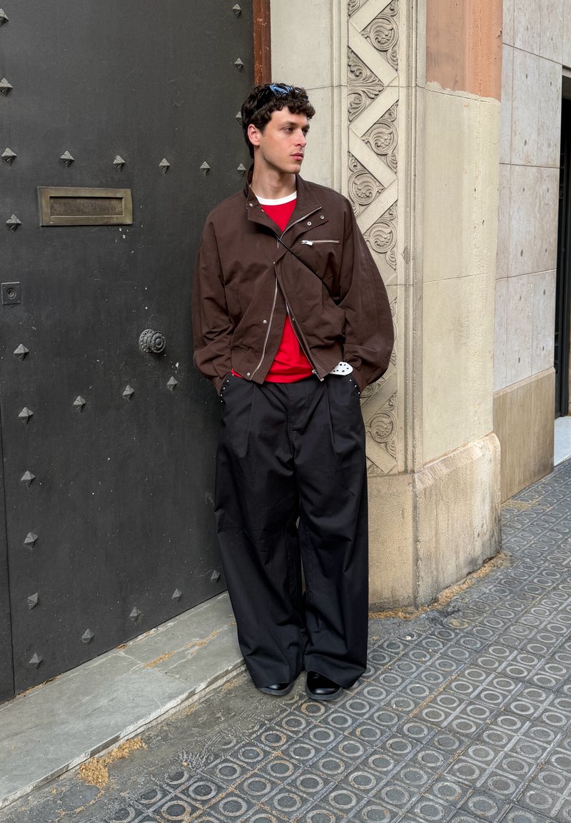 Jeune homme en veste marron et pantalon large noir se tient contre un mur de pierre orné et une porte en métal noir sur le trottoir de la ville.