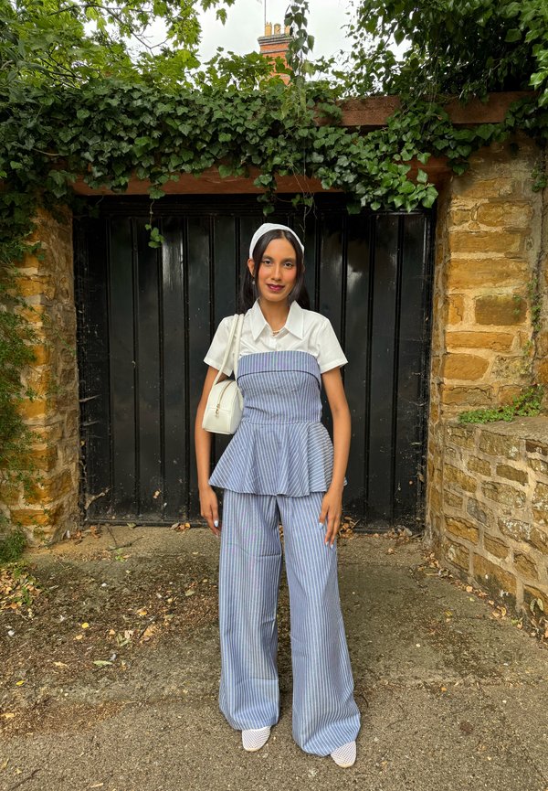 Femme vêtue d'une chemise blanche à manches courtes et d'un pantalon rayé bleu, debout devant une porte noire encadrée de lierre et de murs en pierre. Elle porte un sac blanc.