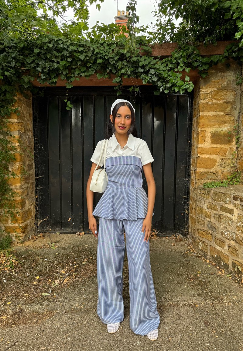 Femme en chemise blanche à manches courtes et pantalon bleu à rayures, debout devant un portail noir encadré de lierre et de murs en pierre. Elle porte un sac blanc.