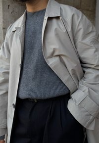 Μπεζ εξωτερικό παλτό πάνω από γκρί ριμπ πουλόβερ, συνδυασμένο με σκούρες παντελόνες. Το παλτό διαθέτει μεγάλους γάντζους και χαλαρή εφαρμογή.