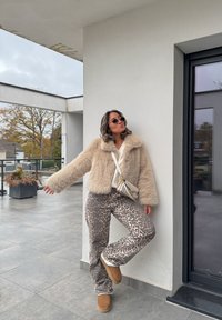 Beige faux fur jacket, white jumper, animal print trousers, brown boots, and a small light handbag; standing against a modern building background.