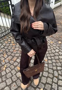 Femme aux longs cheveux bruns portant une veste en cuir noire et un pantalon foncé, tenant un sac à main marron, debout sur un pavé avec des feuilles d'automne.