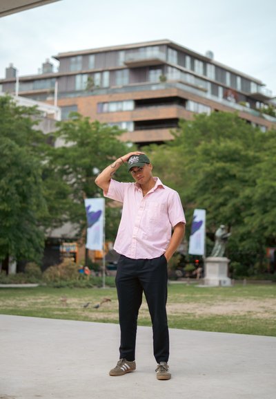 Hombre con una camisa de manga corta rosa claro y pantalones negros, de pie de forma casual. Lleva una gorra verde y zapatillas marrones con rayas blancas.