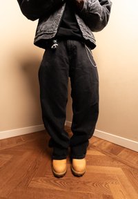 Personne portant un jean noir ample, des bottes jaunes, une veste sombre et un porte-clés accroché au jean, debout sur un sol en bois.