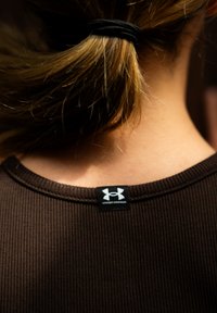 Close-up van de rug van een persoon met blond haar dat vastgebonden is met een zwarte haarband, in een bruine ribbeltrui met een Under Armour-label.