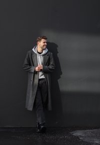 Długi szary wełniany płaszcz na szarym hoodie, czarne dżinsy i czarne buty. Osoba stoi przy ciemnej ścianie, rzucając delikatny cień.