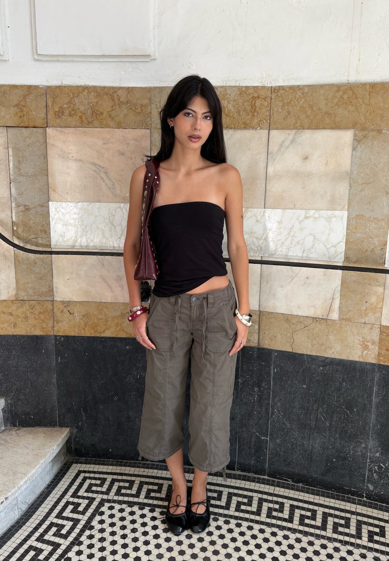 Jeune femme portant un haut tube noir, un pantalon gris ample et des chaussures noires, debout contre un mur carrelé de marbre et un sol à motifs à l'intérieur.