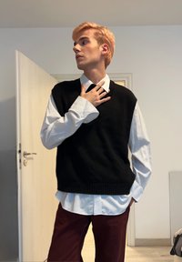 Czarna dzianinowa kamizelka noszona na białej koszuli na guziki z podwiniętymi rękawami, w połączeniu z ciemnymi bordowymi spodniami. Gładka faktura, casualowy, warstwowy wygląd.