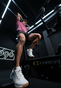 Atlet v růžovém tričku a černých šortkách provádí cvičení na černém fitness stroji. Viditelné bílé sportovní boty, svalnaté nohy a tetování.