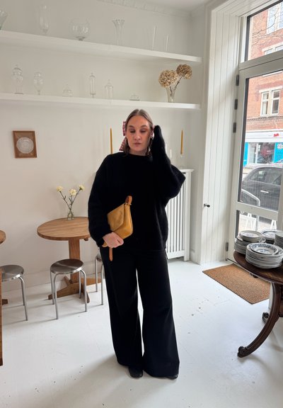 Mujer con atuendo negro sosteniendo un clutch color camel, de pie en una habitación luminosa con mesas de madera, taburetes de metal, estantes con cristalería y una gran ventana.