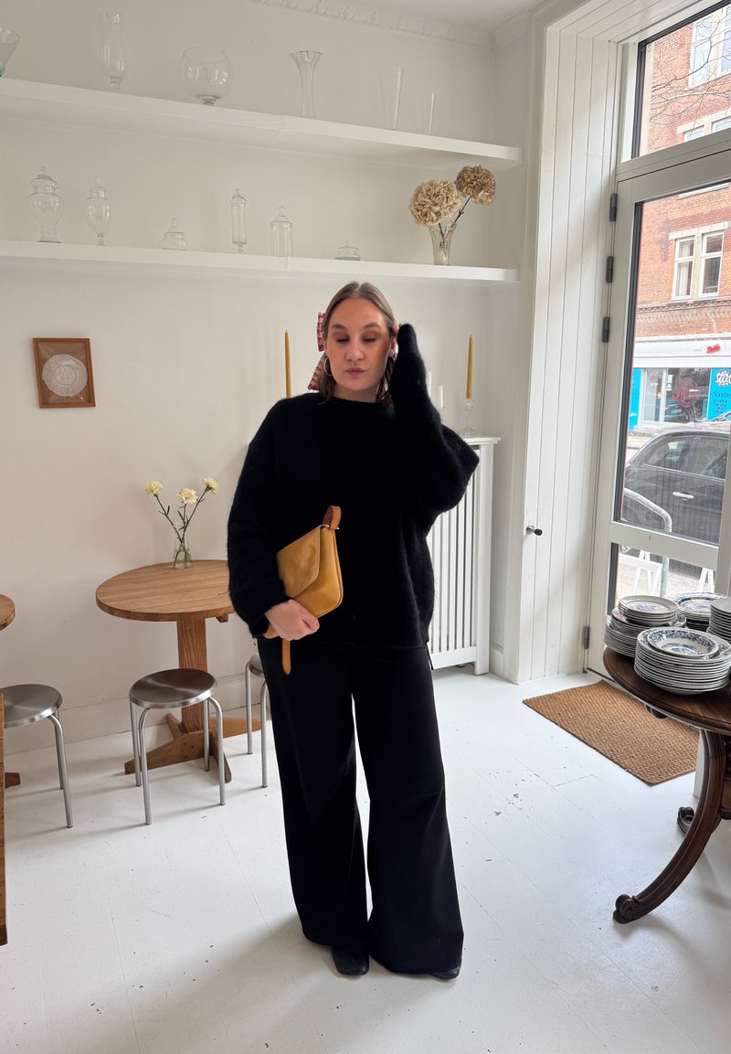 Donna in abito nero che tiene una pochette color cammello, in piedi in una stanza luminosa con tavoli in legno, sgabelli in metallo, scaffali con bicchieri e una grande finestra.